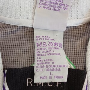 May include: A white and purple sports jersey featuring a tag with the text LAS PRENDAS OFICIALES DEL REAL MADRID ESTAN LICENCIADAS EXCLUSIVAMENTE PARA SU COMERCIALIZACION POR LA MARCA KELME, REF: 96.558, 100% POLYESTER, MADE IN TAIWAN, and R.M.C.F.