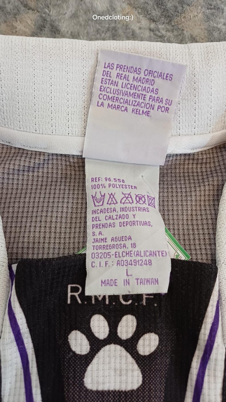 May include: A white and purple sports jersey featuring a tag with the text LAS PRENDAS OFICIALES DEL REAL MADRID ESTAN LICENCIADAS EXCLUSIVAMENTE PARA SU COMERCIALIZACION POR LA MARCA KELME, REF: 96.558, 100% POLYESTER, MADE IN TAIWAN, and R.M.C.F.