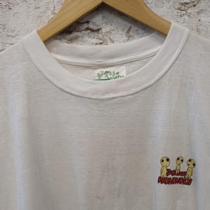 May include: A white short-sleeved t-shirt featuring a small graphic of three yellow Kodama forest spirits from Princess Mononoke. The text Princess Mononoke is printed in red below the figures. A white tag with green Japanese text is at the neck.