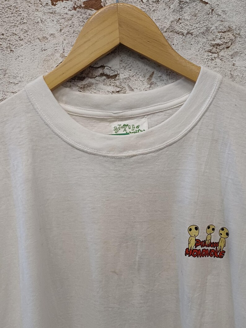 May include: A white short-sleeved t-shirt featuring a small graphic of three yellow Kodama forest spirits from Princess Mononoke. The text Princess Mononoke is printed in red below the figures. A white tag with green Japanese text is at the neck.