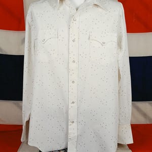 May include: A long-sleeved button-down shirt in off-white featuring a subtle, repeating dark speckle pattern. The shirt includes a pointed collar, two chest pockets with pointed flaps, and a front placket secured with round, light-coloured buttons.