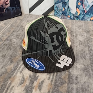 May include: A black and white baseball cap featuring a DC logo, a Ford logo, and the number 43. The cap has a striped pattern, a grey paint splatter design, and a flat brim. It includes small yellow Pirelli branding on the side panels.
