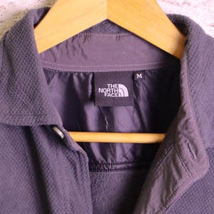 May include: A dark grey button-up jacket with a textured fleece exterior and a smooth interior lining. The garment features a collar, a front button closure, and a black interior label with the text The North Face and the size M clearly visible.