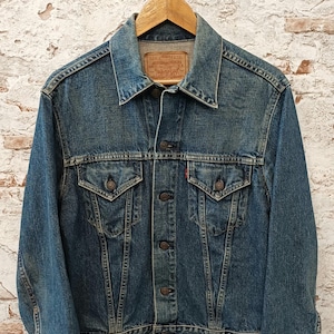May include: A blue denim jacket featuring a pointed collar, long sleeves with buttoned cuffs, and a front button closure. It includes two chest pockets with buttoned flaps, a small red tab on the left pocket, and a tan leather brand label at the neck.