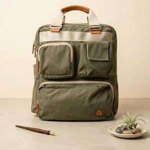 May include: An olive green canvas backpack featuring a large rectangular main compartment, two smaller front pockets, and a wide horizontal beige strap across the top. It includes a tan leather-wrapped handle and silver metal hardware accents.