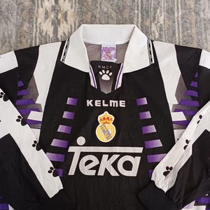 May include: A black, white, and purple long-sleeve soccer jersey featuring the Kelme brand name, the Real Madrid club crest, and the Teka sponsor logo on the front. The shirt has a white collar and black cuffs with a repeating paw print pattern.