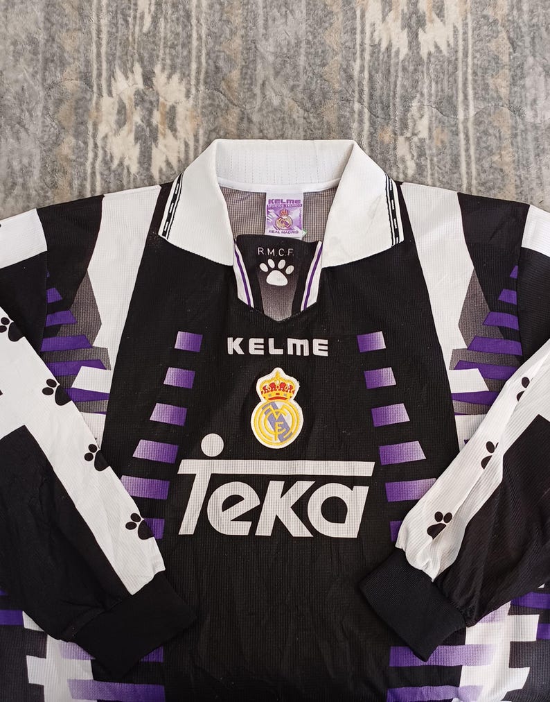 May include: A black, white, and purple long-sleeve soccer jersey featuring the Kelme brand name, the Real Madrid club crest, and the Teka sponsor logo on the front. The shirt has a white collar and black cuffs with a repeating paw print pattern.