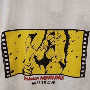 May include: A white t-shirt featuring a rectangular yellow graphic designed like a film strip. The graphic depicts two animated characters in black ink, with the red and black text "Princess MONONOKE WILL TO LIVE" printed below the illustration.