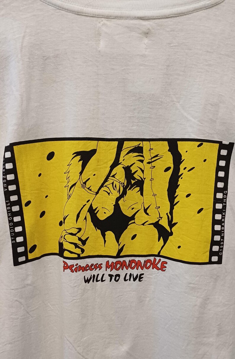 May include: A white t-shirt featuring a rectangular yellow graphic designed like a film strip. The graphic depicts two animated characters in black ink, with the red and black text "Princess MONONOKE WILL TO LIVE" printed below the illustration.