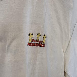 May include: A white short-sleeved t-shirt featuring a small graphic on the chest. The graphic displays three yellow Kodama figures from Princess Mononoke above the red text Princess MONONOKE. The shirt has a crew neckline and a plain white fabric.