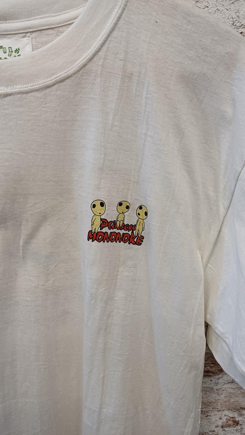 May include: A white short-sleeved t-shirt featuring a small graphic on the chest. The graphic displays three yellow Kodama figures from Princess Mononoke above the red text Princess MONONOKE. The shirt has a crew neckline and a plain white fabric.