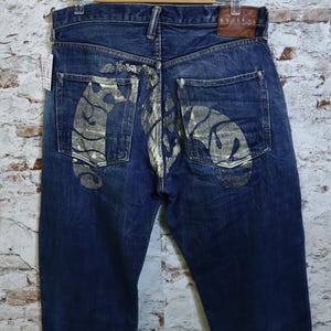 Puede incluir: Vaqueros de mezclilla azul oscuro con un diseño gráfico pintado en dorado metálico en los bolsillos traseros y el asiento. Los pantalones incluyen un parche de cuero marrón en la cintura trasera, bragueta de botones y cinco bolsillos.