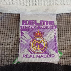 May include: A white clothing tag featuring the purple Kelme logo, the text Sponsor Tecnico, and the Real Madrid crest with a crown. Below this, a black tag displays the white text R.M.C.F. The background is a textured grey and purple fabric.