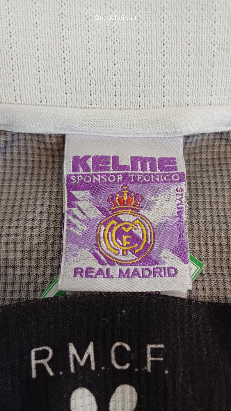 May include: A white clothing tag featuring the purple Kelme logo, the text Sponsor Tecnico, and the Real Madrid crest with a crown. Below this, a black tag displays the white text R.M.C.F. The background is a textured grey and purple fabric.