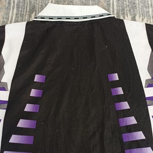 May include: A black, white, and purple collared shirt featuring a textured fabric. The design includes a white collar with a thin black trim, a solid black center panel, and vertical purple gradient geometric patterns on the sides of the garment.