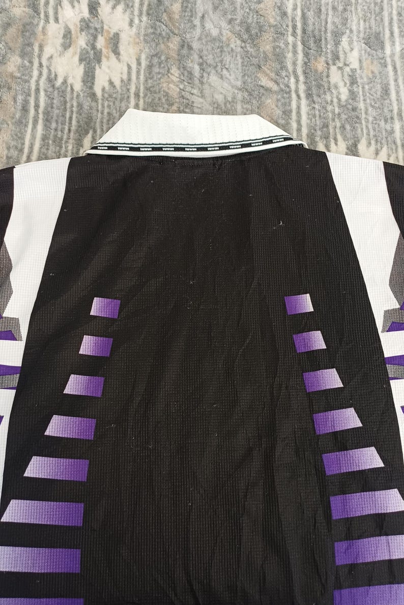 May include: A black, white, and purple collared shirt featuring a textured fabric. The design includes a white collar with a thin black trim, a solid black center panel, and vertical purple gradient geometric patterns on the sides of the garment.