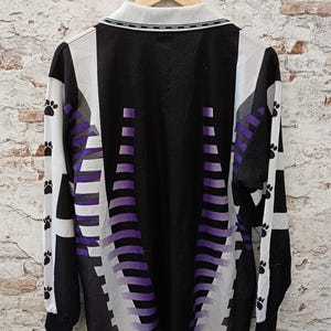 May include: A long-sleeve shirt featuring a black base with white side panels and a purple gradient pattern. The sleeves are accented with a repeating black paw print design. The shirt includes a white collar with a black trim detail.