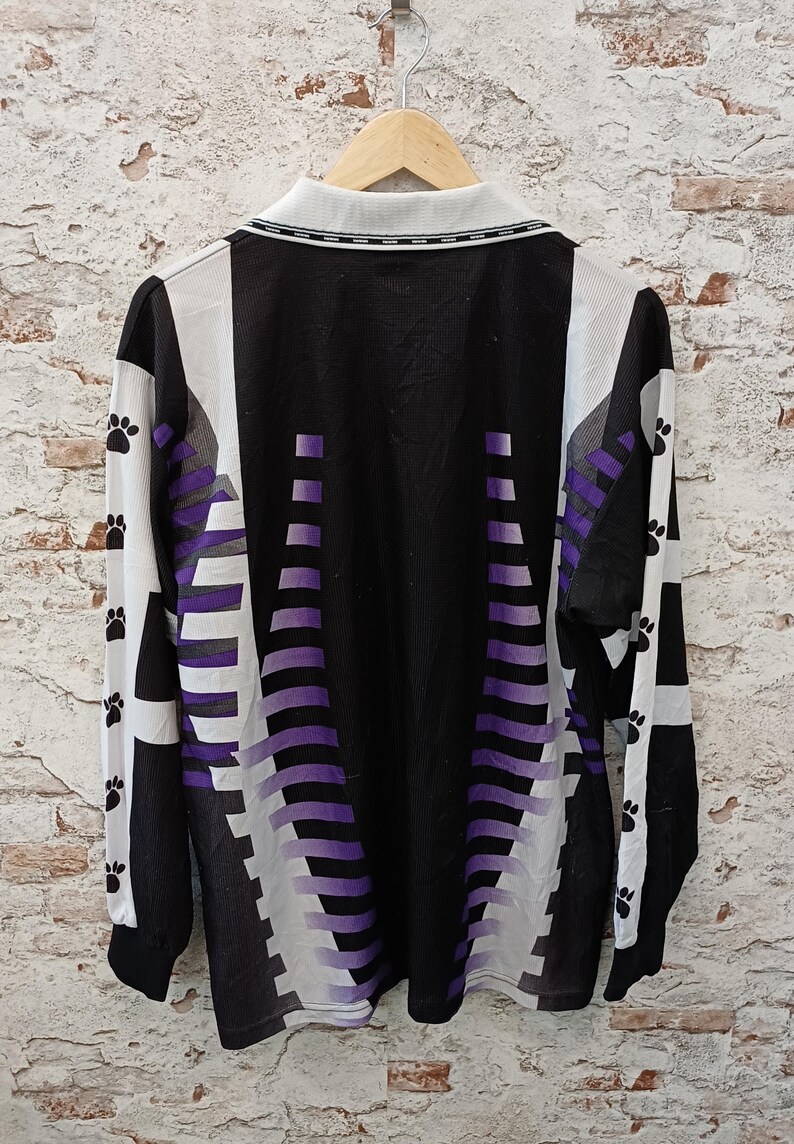 May include: A long-sleeve shirt featuring a black base with white side panels and a purple gradient pattern. The sleeves are accented with a repeating black paw print design. The shirt includes a white collar with a black trim detail.