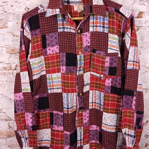 May include: A long-sleeved button-down shirt featuring a patchwork design of various red, pink, and white plaid and floral fabric squares. The shirt includes a collar, a single chest pocket, and light-coloured buttons. The label reads Candy Fantasy.