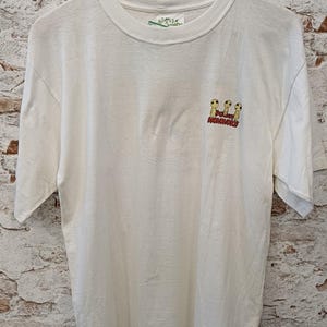 May include: A white short-sleeve t-shirt featuring a small graphic on the upper right chest. The graphic depicts three yellow Kodama figures from Princess Mononoke with the red text Princess Mononoke printed directly underneath the figures.