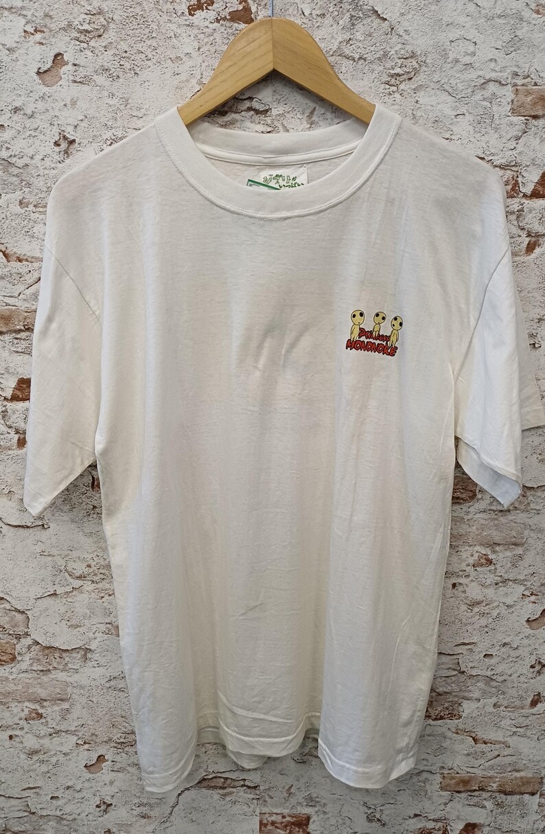 May include: A white short-sleeve t-shirt featuring a small graphic on the upper right chest. The graphic depicts three yellow Kodama figures from Princess Mononoke with the red text Princess Mononoke printed directly underneath the figures.