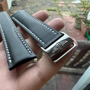 Könnte beinhalten: Schwarzes Leder-Uhrenarmband mit weißen Nähten. Das Armband hat eine silberfarbene Schließe mit einem geflügelten Emblem. Das Uhrenarmband ist für einen klassischen und raffinierten Look konzipiert.