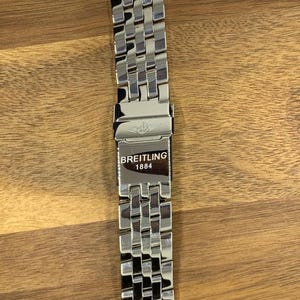 Könnte beinhalten: Ein silberfarbenes Breitling-Uhrenarmband mit einem gewebten Gliederdesign. Der Verschluss zeigt das Breitling-Logo und den Text "BREITLING 1884". Das Armband wird auf einer Holzoberfläche präsentiert.