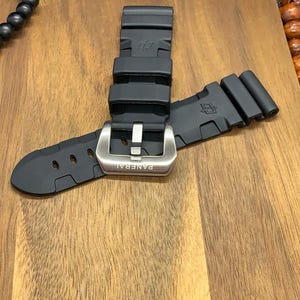 May include: A black rubber watch strap with a silver buckle. The buckle is engraved with the word "PANERAI". The strap is lying on a wooden surface.