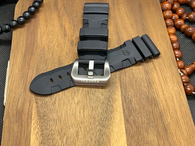 May include: A black rubber watch strap with a silver buckle. The buckle is engraved with the word "PANERAI". The strap is lying on a wooden surface.
