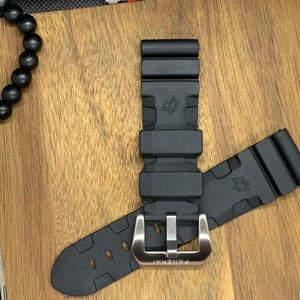 May include: Black rubber watch strap with a silver buckle. The strap is lying on a wooden surface. The buckle is engraved with the word "PANERAI".