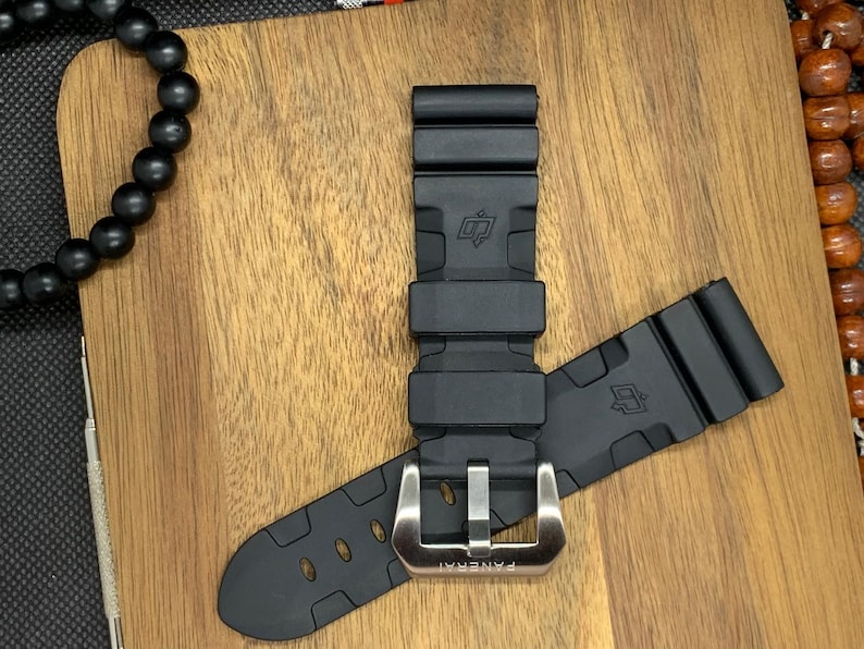 May include: Black rubber watch strap with a silver buckle. The strap is lying on a wooden surface. The buckle is engraved with the word "PANERAI".