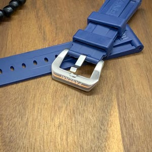 May include: A navy blue watch strap with a silver buckle. The strap is made of a flexible material and has several holes for adjustment. The buckle is engraved with the word "PANERAI".