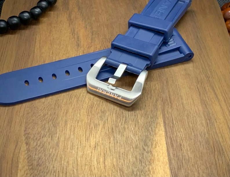May include: A navy blue watch strap with a silver buckle. The strap is made of a flexible material and has several holes for adjustment. The buckle is engraved with the word "PANERAI".