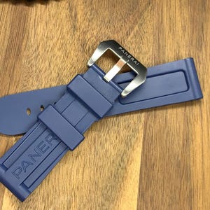 May include: A navy blue watch strap with a silver buckle. The strap is made of a flexible material and features the brand name "PANERAI" embossed on the band. The buckle is rectangular with the brand name engraved.