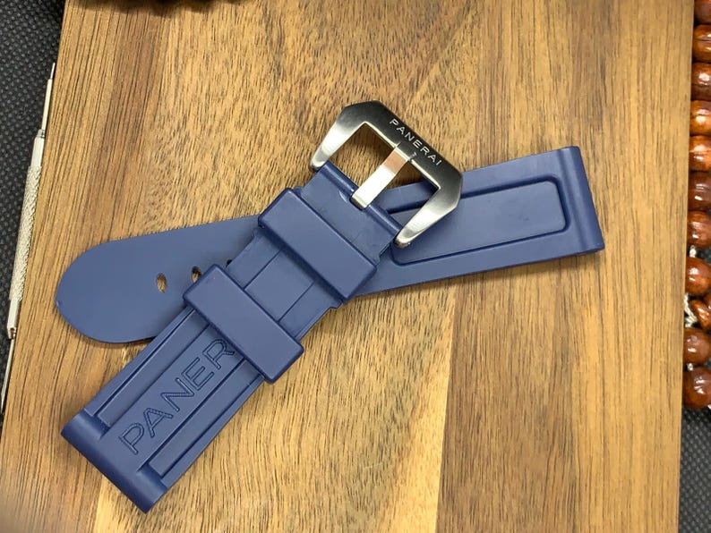 May include: A navy blue watch strap with a silver buckle. The strap is made of a flexible material and features the brand name "PANERAI" embossed on the band. The buckle is rectangular with the brand name engraved.