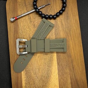 May include: An olive green watch strap with the words "OFFICINE" and "PANER" embossed on it. The strap has a silver buckle and three holes. A black beaded bracelet and a small screwdriver are also in the image.