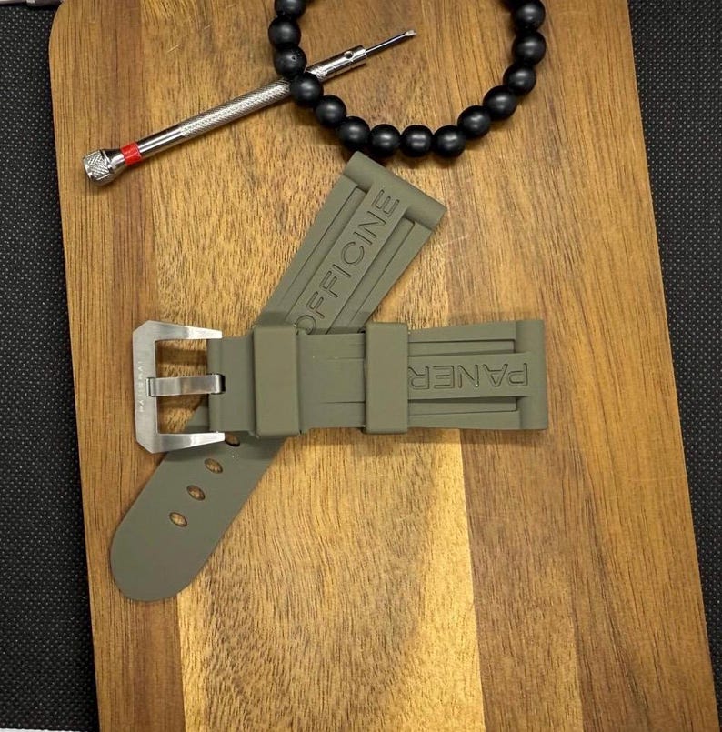 May include: An olive green watch strap with the words "OFFICINE" and "PANER" embossed on it. The strap has a silver buckle and three holes. A black beaded bracelet and a small screwdriver are also in the image.
