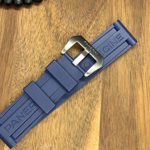 May include: A navy blue watch strap with the brand name "PANERAI" embossed on the strap. The strap has a silver-colored buckle with the brand name engraved. The strap is made of a flexible material.