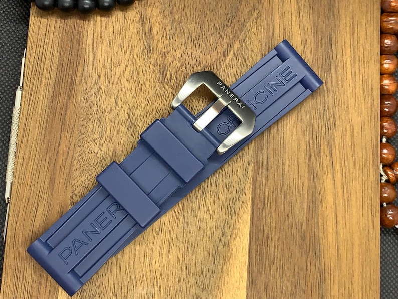 May include: A navy blue watch strap with the brand name "PANERAI" embossed on the strap. The strap has a silver-colored buckle with the brand name engraved. The strap is made of a flexible material.