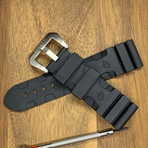 May include: Black watch strap with a silver buckle, featuring the brand name "Panerai". The strap is made of a flexible material and has a textured design. A small screwdriver is visible near the strap.