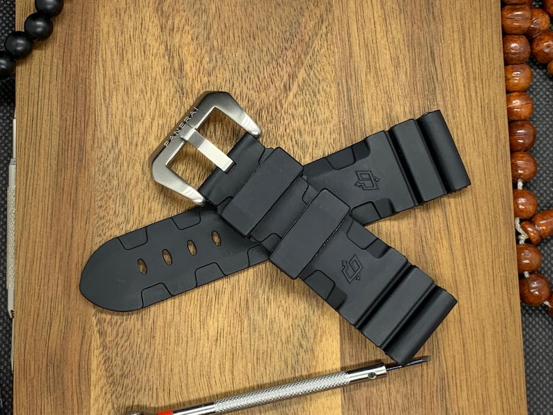 May include: Black watch strap with a silver buckle, featuring the brand name "Panerai". The strap is made of a flexible material and has a textured design. A small screwdriver is visible near the strap.