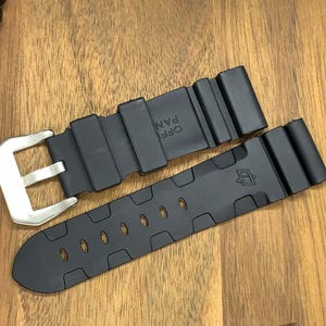 May include: Black watch strap with a silver buckle. The strap is made of a flexible material and features a series of oval cutouts. The watch strap is shown on a wooden surface.