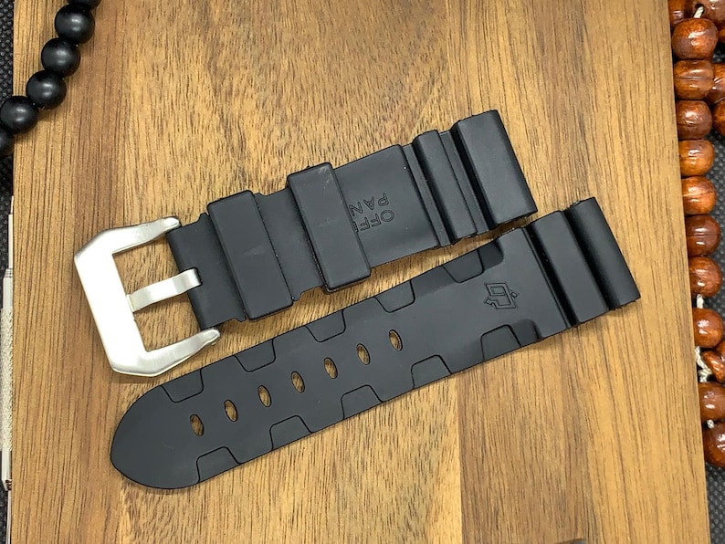 May include: Black watch strap with a silver buckle. The strap is made of a flexible material and features a series of oval cutouts. The watch strap is shown on a wooden surface.