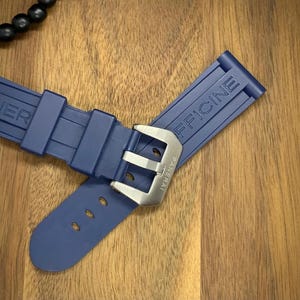 May include: A navy blue watch strap with the brand name "PANERAI OFFICINE" embossed on the rubber. The strap features a silver-colored buckle and is displayed on a wooden surface.