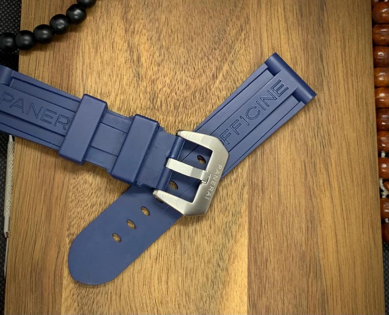 May include: A navy blue watch strap with the brand name "PANERAI OFFICINE" embossed on the rubber. The strap features a silver-colored buckle and is displayed on a wooden surface.