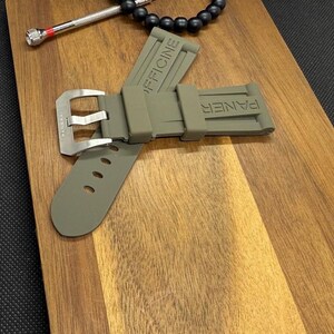 May include: An olive green watch strap with a silver buckle, embossed with the words "PANER" and "OFFICINE". A small silver tool and a black beaded bracelet are also visible.