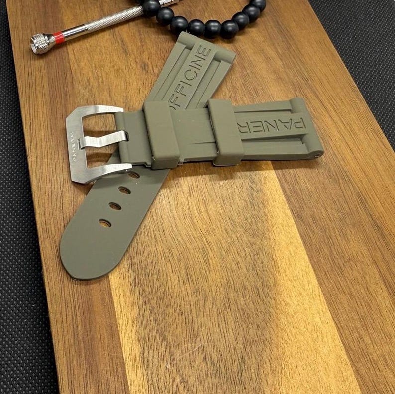 May include: An olive green watch strap with a silver buckle, embossed with the words "PANER" and "OFFICINE". A small silver tool and a black beaded bracelet are also visible.