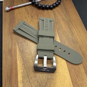 May include: An olive green watch strap with the word "PANERAI" embossed on the strap and buckle. The strap is made of a flexible material and has multiple holes for adjustment. The buckle is silver in colour.