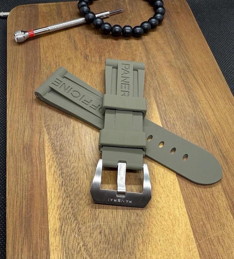 May include: An olive green watch strap with the word "PANERAI" embossed on the strap and buckle. The strap is made of a flexible material and has multiple holes for adjustment. The buckle is silver in colour.