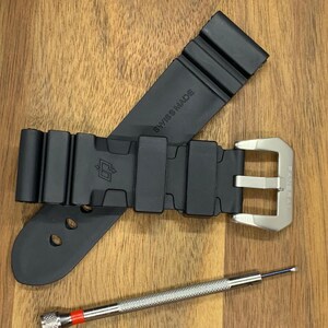 May include: Black rubber watch strap with a silver buckle and a small screwdriver. The strap is marked "SWISS MADE". The watch strap is lying on a wooden surface.